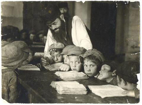 A group of elementary school students look at their notebooks as a teacher helps one of them. Circa 1920s Poland. 