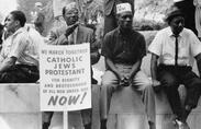 Photograph shows some participants in the civil rights march sitting on a wall resting, one holds a placard which reads, "We march together, Catholics, Jews, Protestant, for dignity and brotherhood of all men under God, Now!" Image used in Reconstruction video series.