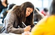 A student in a grey sweatshirt looks down at a paper with a pencil in hand.