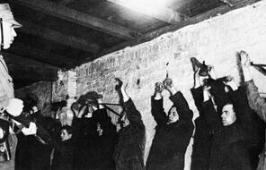 A man in uniform stands before a line of men and women against a brick wall with their hands up. 
