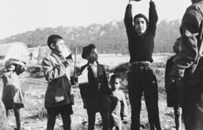 A group of boys gather in the Los Arenas camp. One boy stands in the middle holding a rock over his head while others look at him.