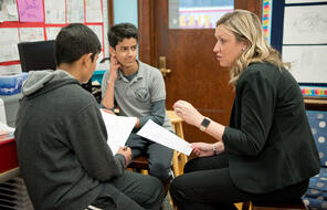 A teacher discusses a handout with two students seated at a classroom table