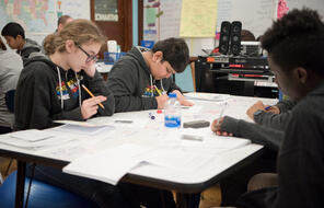 Three students sit around a shared desk in their classroom and work on a handout.