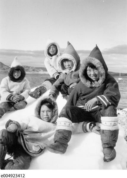 Young boys sitting on snow wearing hooded coats lined with fur and boots. 