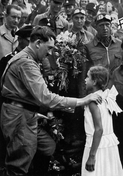 Adolf Hitler has his hand placed on the soldier of a young girl in a white dress. 