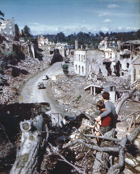 Two boys sit on a branch in the foreground looking out at destroyed buildings and countryside. 