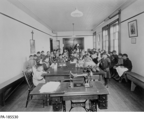  Children in a classroom with a sewing machine on a table. A nun stands in the back of the classroom. Taken circa 1929.