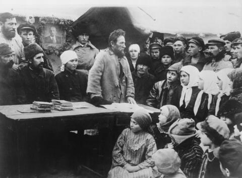 A bearded man stands at a table, surrounded by a listening crowd of peasants
