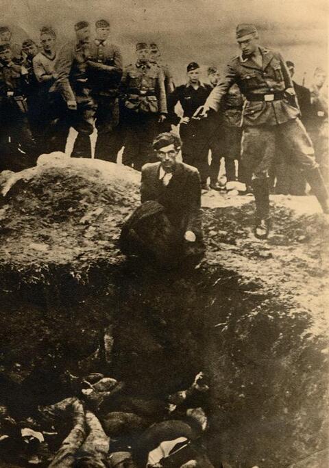 Nazi soldier holds gun to a man's head who is kneeled next to a ditch full of bodies. 