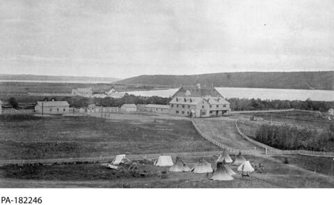 A rural setting with teepees in the foreground and a group of buildings in the background.