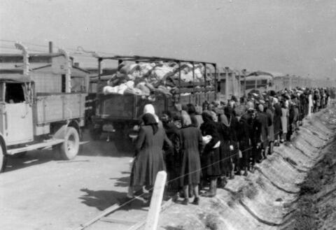 Photograph of a trailer full of clothes and other belongings of new camp prisoners with a group of women on the side of the road.