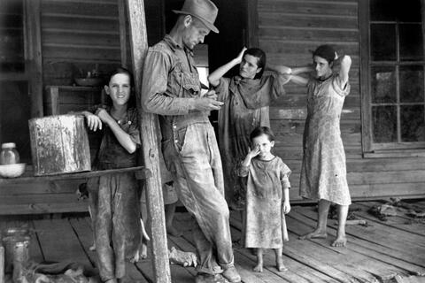 A man and four girls stand on a porch, wearing torn and dirty clothing.