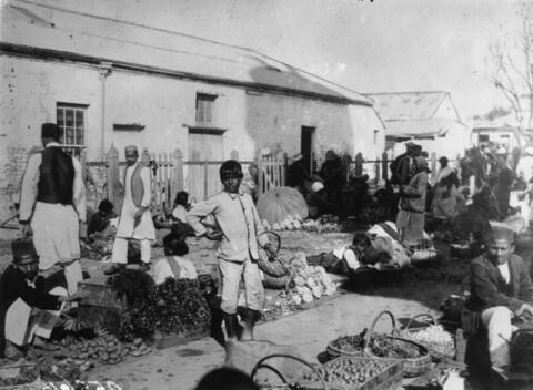 An uncovered outdoor fruit market with baskets of fruit on the ground and men, women, and children buying and selling fruit.