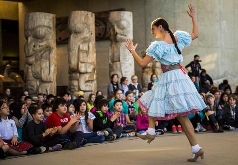 A woman in a blue dress with a colorful waist-sash is dancing for a group of children.