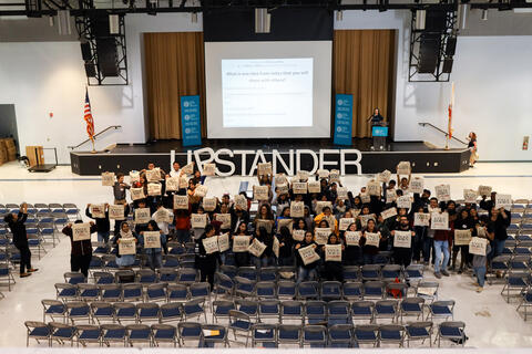 A large group of students pose for an picture with the word "Upstander" in the background.