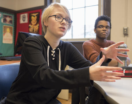 Two students sitting in a classroom with one student talking and gesturing