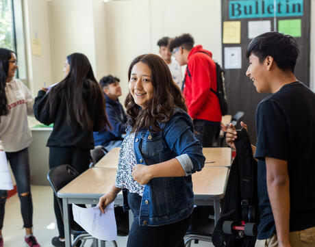 Students move around the classroom in conversation with each other. One student looks directly into the camera with a smile on their face.