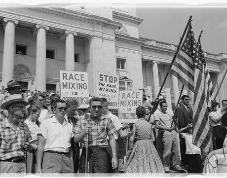 A group of people, several holding signs and American flags, protesting the admission of the "Little Rock Nine" to Central High School.
