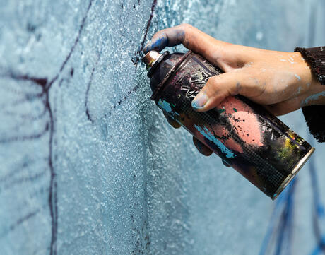 A young man holds a dark can of spray paint close to a blue wall to sketch his graffiti