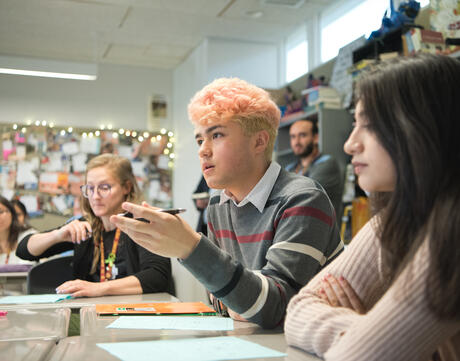 A student with pink hair speaks in a classroom with their hand raised.