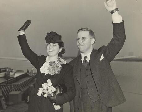 Martha and Waitstill Sharp wave to a crowd before leaving New York City for Europe. Martha wears a corsage of flowers on her coat and holds a bouquet of flowers in her left hand. Stamped in ink on verso: "Photo by William T. Hoff, New York Municipal Airport"