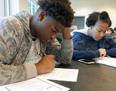 Two Black students write at their desks. 